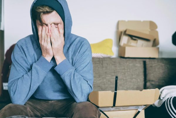 En person i blå luvtröja sitter i en soffa med ansiktet i händerna och ser stressad ut. Framför dem står en öppen kartong och några föremål på bordet, och fler lådor i bakgrunden.