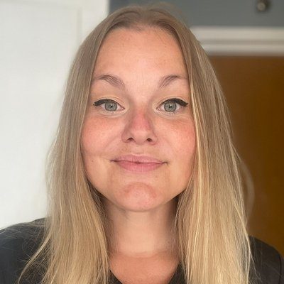 En kvinna med långt blont hår och ljusa ögon ler mot kameran. Hon bär svart eyeliner och en svart topp och står inomhus med en vit dörr och en trädörr i bakgrunden.
